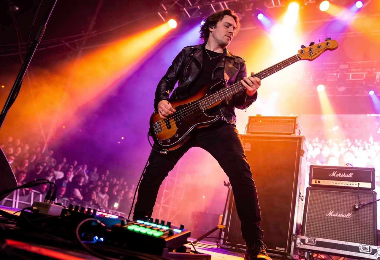 Bassist spielt E-Bass live auf einer Konzertbühne mit farbiger Bühnenbeleuchtung und Verstärkern im Hintergrund