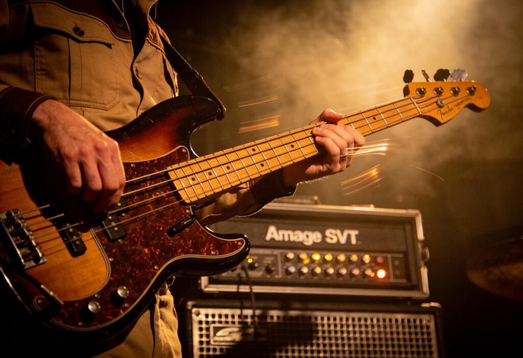 Ein Bassist in einem khakifarbenen Hemd, der einen Bass in einer live Musik-Performance spielt.