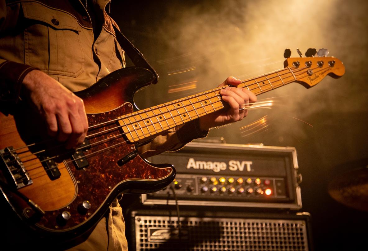 Ein Bassist in einem khakifarbenen Hemd, der einen Bass in einer live Musik-Performance spielt.