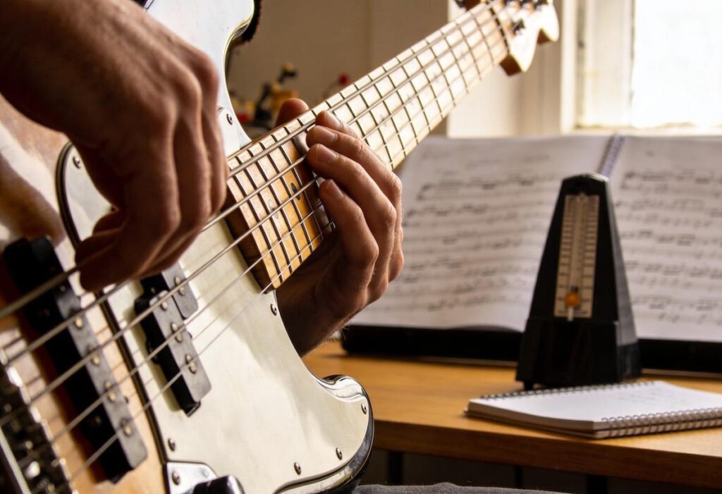 Ein Musiker spielt Bassgitarre, während Noten und ein Metronom auf dem Tisch stehen.