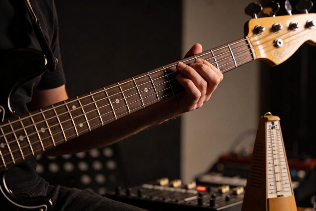 Ein Bassist spielt eine E-Bassgitarre mit einer Hand auf den Saiten und einer Metronom im Vordergrund.